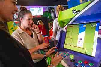 Two women playing on an arcade machine with TVs showing people playing VR games behind them in VRcafe Haarlem during a friends outing