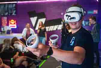 Man in navy T-shirt playing a shooter VR game in front of a purple wall with big VRcafe logo on it during a company outing