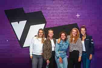 A group of women posing against a purple wall with a big VRcafe logo on it after their bachelor's party outing
