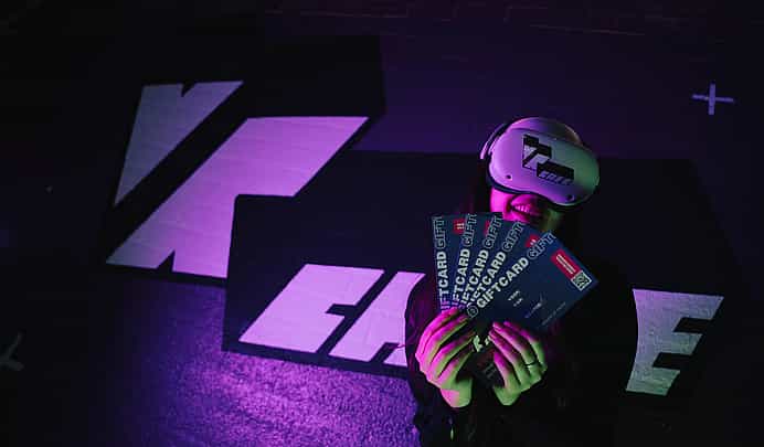 Woman in Meta Quest 2 headset holding 5 giftcards standing against a wall with a big VRcafe Haarlem logo on it
