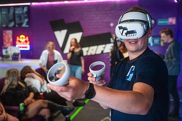 Man in navy T-shirt playing a shooter VR game in front of a purple wall with big VRcafe logo on it during a company outing