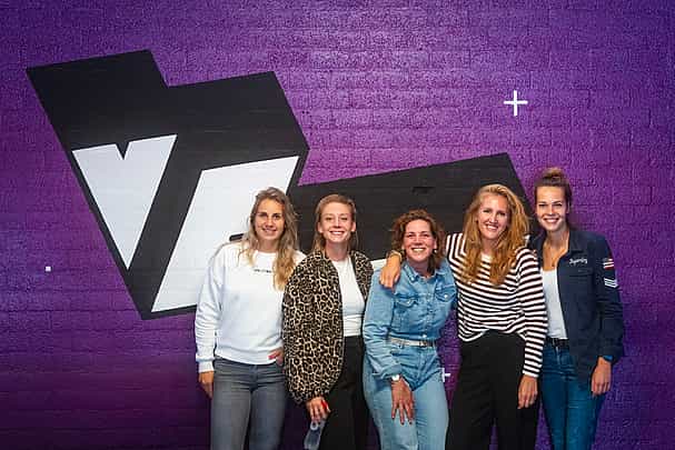 A group of women posing against a purple wall with a big VRcafe logo on it after their bachelor's party outing