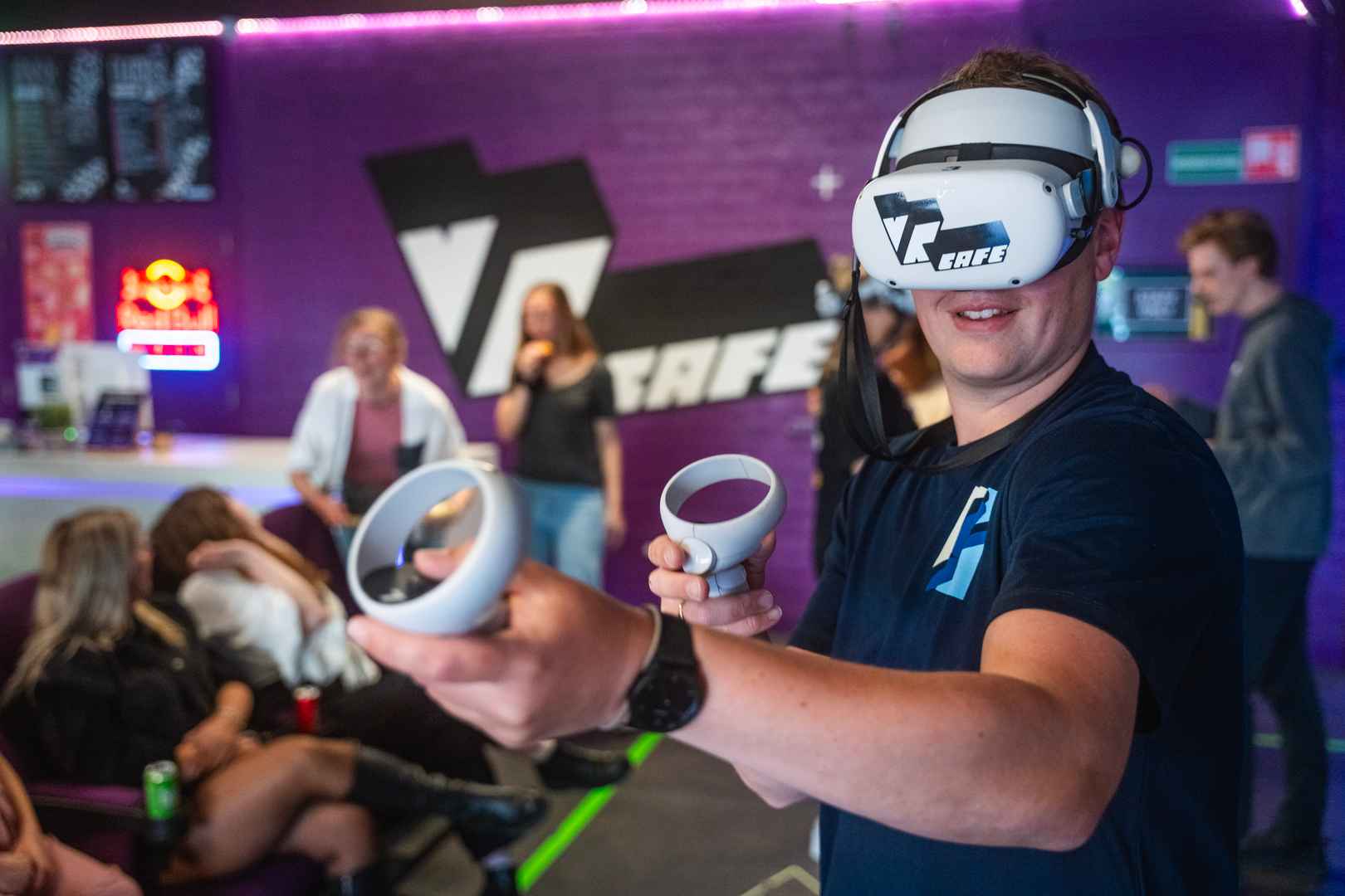 Man in navy T-shirt playing a shooter VR game in front of a purple wall with big VRcafe logo on it during a company outing